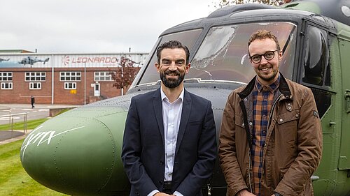 Adam Dance MP and Ben Clarke from Unite stand together in front of a military helicopter at the Leonardo facility. Adam wears a brown jacket and plaid shirt, while Ben is dressed in a navy suit with a white shirt. The Leonardo logo is visible on the building in the background.