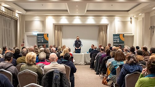 Adam Dance MP stands at the front of a well-attended public meeting, addressing a seated audience in a conference room. Behind him, a panel of individuals is seated at a table, and two banners read "MEET YOUR MP - ADAM DANCE." The room is filled with people listening attentively.