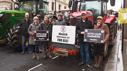 A group of farmers and supporters stand in front of large tractors on a city street, holding signs that read "WITH OUR FARMERS #together" and a banner stating "Abolish Inheritance Tax FOR ALL!" The individuals are dressed in outdoor work clothing, including jackets, hats, and boots. 