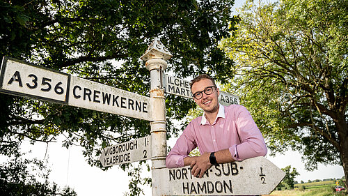 Adam Dance, MP for Yeovil, smiling and leaning against a vintage signpost at a rural crossroads. The signpost displays directions to Crewkerne, Ilchester, Martock, and North Perrott, set against a sunny countryside backdrop with trees and open fields.