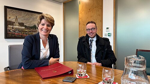 Adam Dance MP and Emma Hardy MP sit at a wooden table in a meeting room, both smiling. Emma Hardy wears a navy blazer and has a red folder in front of her. Adam Dance wears a black coat, white shirt, and patterned tie. The table has glasses of water, a jug, and a remote control. Behind them, a painting of a royal event hangs on the wall. The room has wooden and glass doors.