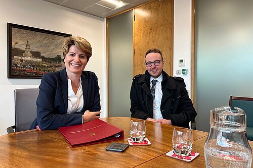 Adam Dance MP and Emma Hardy MP sit at a wooden table in a meeting room, both smiling. Emma Hardy wears a navy blazer and has a red folder in front of her. Adam Dance wears a black coat, white shirt, and patterned tie. The table has glasses of water, a jug, and a remote control. Behind them, a painting of a royal event hangs on the wall. The room has wooden and glass doors.