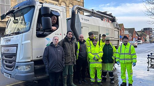 A group of workers in high-visibility yellow jackets stand together on a wet pavement next to a large street-cleaning vehicle with 'Somerset' branding. Adam Dance wears a dark jacket and green wellington boots and is standing among them, while a driver leans out of the truck’s window. The background features historic buildings and a town street with cars parked along the road.