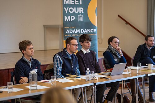 Adam Dance, MP for Yeovil, sits alongside other participants at a table during his farming forum. Behind him, a banner reads 'Meet Your MP – Adam Dance, MP for Yeovil and South Somerset.' The setting is a community hall, and documents, water glasses, and a laptop are visible on the table.