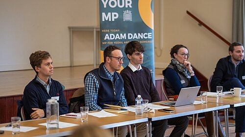 Adam Dance, MP for Yeovil, sits alongside other participants at a table during his farming forum. Behind him, a banner reads 'Meet Your MP – Adam Dance, MP for Yeovil and South Somerset.' The setting is a community hall, and documents, water glasses, and a laptop are visible on the table.