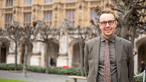 Adam Dance MP standing outdoors in front of the historic architecture of the Houses of Parliament. He is wearing a grey suit, a patterned tie, and glasses, smiling at the camera. The background features leafless trees and a well-maintained garden area.