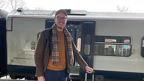 Adam Dance, MP for Yeovil, smiling on a train platform, holding a grey suitcase with a train in the background. He is dressed casually in a brown jacket, scarf, and cap, ready for travel.