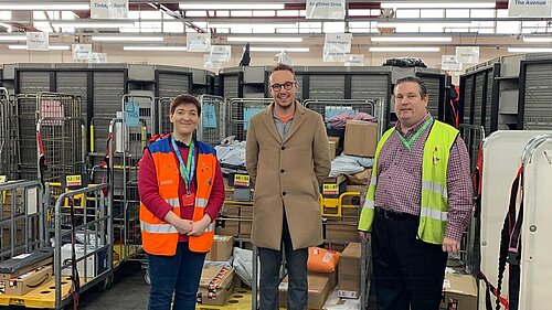 Adam Dance MP standing in a Royal Mail sorting facility alongside two staff members. He is wearing a beige coat and glasses, while the staff wear high-visibility vests, one orange and one yellow. They are surrounded by trolleys filled with parcels and letters, with visible signage and red beams in the background.